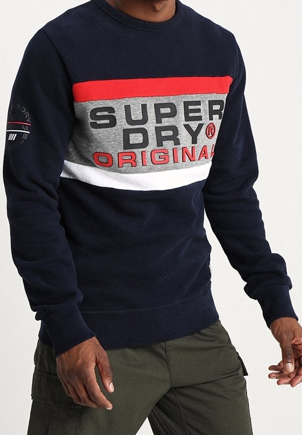 Uomo che indossa un felpa blu navy con strisce rosse, grigie e bianche e la scritta "SUPERDRY ORIGINAL", abbinata a pantaloni cargo verde scuro.