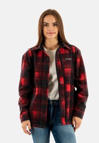 Camisa de franela a cuadros en rojo y negro con botones a presión, mangas largas, collar y un parche de logo en el lado izquierdo del pecho. Llevada sobre una camisa clara.