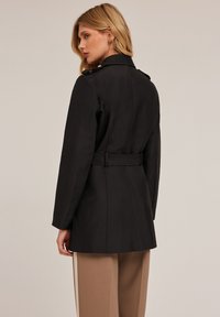 Abrigo negro ajustado con cinturón, que presenta un cuello estructurado y mangas largas. Tejido liso sin patrones visibles ni detalles de hardware.