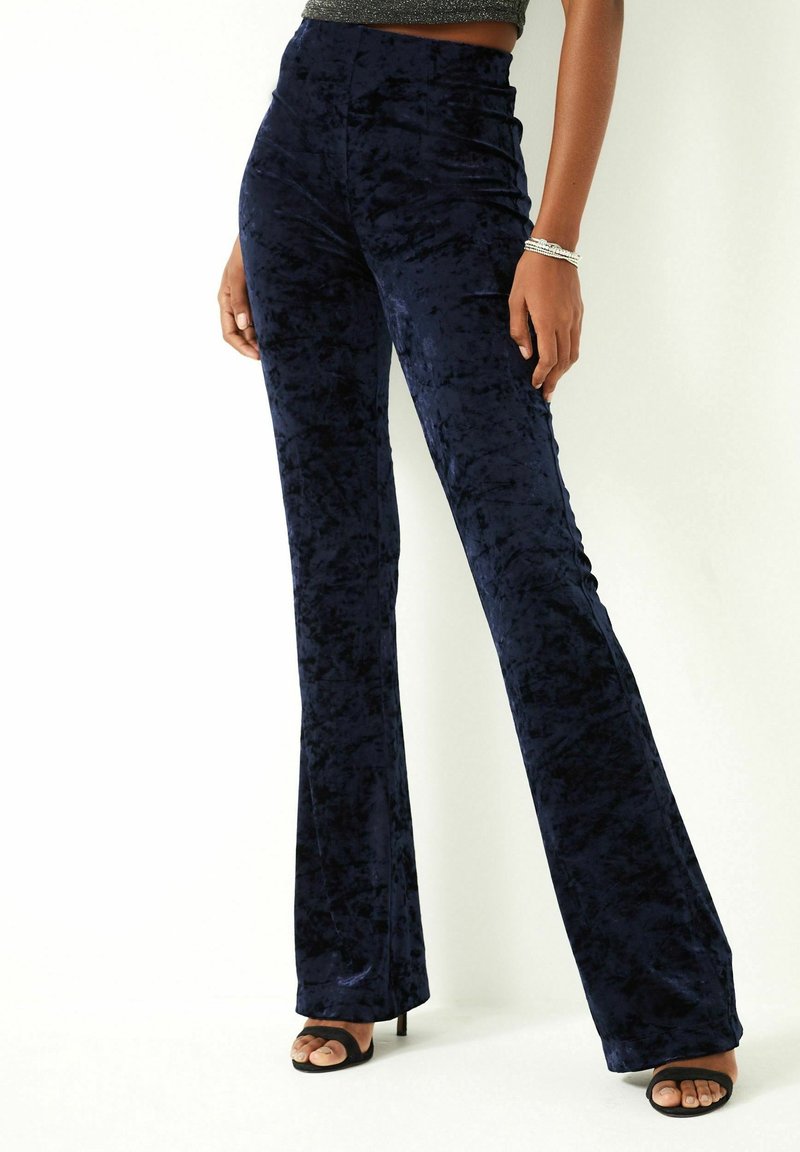 Femme portant un pantalon évasé en velours bleu foncé taille haute et des talons ouverts noirs, debout devant un fond blanc.