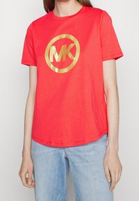 Woman wearing a bright red short-sleeve t-shirt with a large gold "MK" logo and light blue denim jeans.