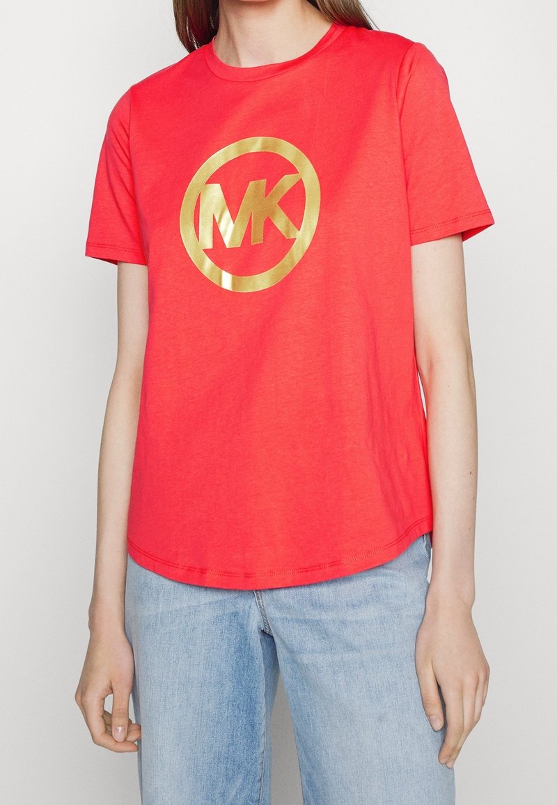 Woman wearing a bright red short-sleeve t-shirt with a large gold "MK" logo and light blue denim jeans.