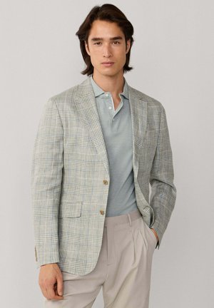 Man draagt een lichtgrijs geruit colbert over een gedempte groene polo en beige geplooide broek, staat met één hand in de zak tegen een effen achtergrond.