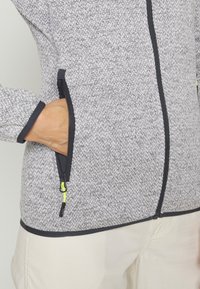 Grå tekstureret zip-jakke med sort kant og lommer, med neonlys detaljer på lynlåse. Nærbillede af hånd i lommen.