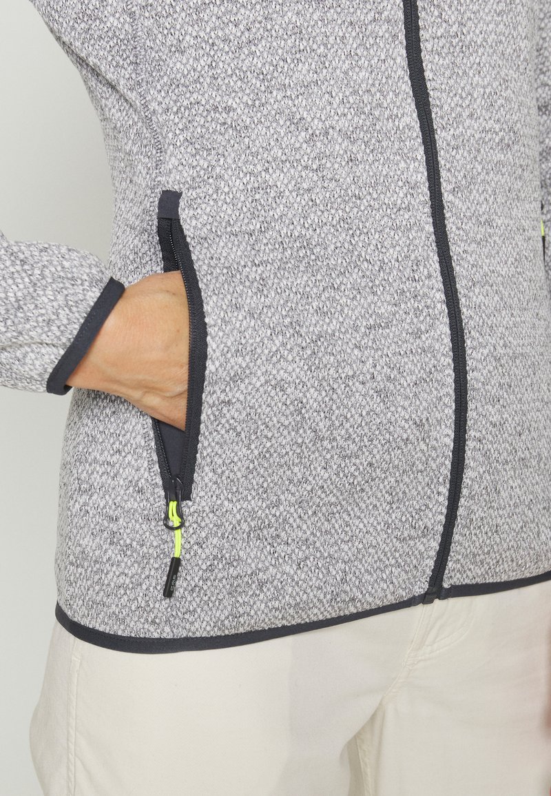 Giacca grigia con texture, con cerniera e rifiniture nere e tasche, caratterizzata da dettagli neon sulle cerniere. Primo piano della mano nei dettagli della tasca.