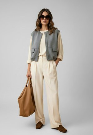 Vrouw met zonnebril, crèmekleurige blouse en broek, grijze mouwloze vest, bruine loafers, houdt grote bruine draagtas vast, staand tegen een grijze achtergrond.