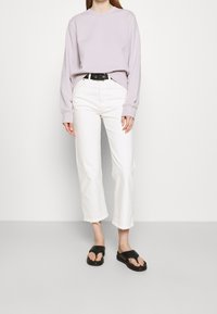 Sudadera lila clara, pantalones vaqueros blancos cortados con dobladillo deshilachado, cinturón negro y chanclas negras. Estilo casual, ajuste relajado.