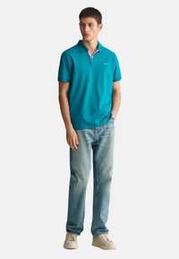 Türkiz pólóing, pink színű gallérral, világoskék farmerral és bézs sportcipővel viselve. Slim fit szabású és rövid ujjú.