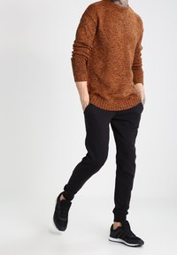 Pull en tricot texturé brun, coupe décontractée, associé à un pantalon de survêtement noir et des baskets noires ; cette tenue met l'accent sur le confort et le style décontracté.