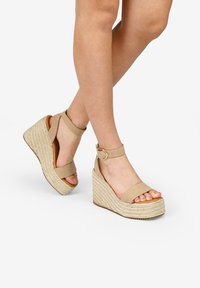 Sandales compensées beiges avec une semelle en jute texturé, deux larges brides au niveau des orteils et de la cheville, et un accent en boucle métallique.