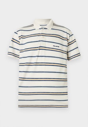 Kremo spalvos piqué polo marškinėliai su horizontaliomis mėlynomis ir oranžinėmis juostelėmis. Yra apykaklė, trijų sagų sijonėlis ir siuvinėtas logotipas.