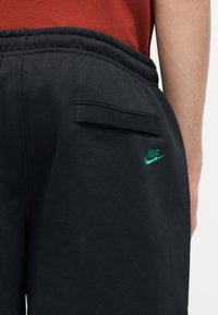 Κοντινό πλάνο μαύρου παντελονιού φόρμας Nike που δείχνει μια πίσω τσέπη με καπάκι και πράσινο κεντημένο λογότυπο Nike στον γοφό, φορεμένο με κόκκινη μπλούζα.