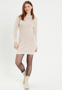 Vestido de punto en un color beige claro, con mangas largas y textura acanalada. Presenta botones decorativos en los hombros y una silueta ajustada.