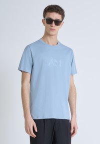 Camiseta azul claro de manga corta con cuello redondo y logo en tonalidad en el pecho. La tela es suave y ligeramente elástica.
