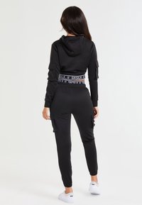 Chándal negro que incluye una sudadera corta con capucha y bolsillos laterales, combinada con joggers ajustados. Destacado con una cinturilla elástica de marca.