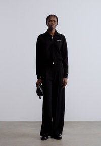 Fekete cipzáras dzseki diszkrét logóval, széles szárú fekete nadrággal párosítva. Kiegészítők között egy kis fekete táska. Minimalista dizájn.