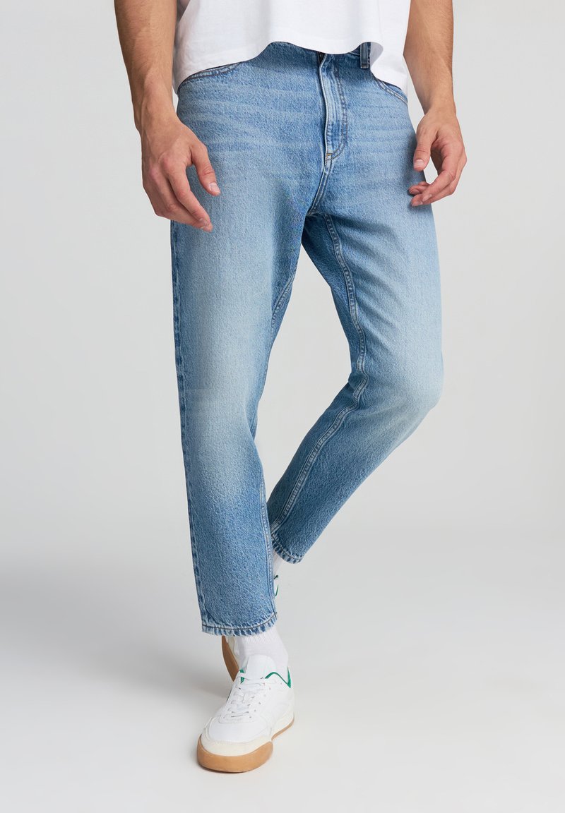Jeans in denim azzurro sbiadito con taglio dritto, caratterizzati da leggere sfumature e design classico a cinque tasche. Abbinati a sneakers bianche.