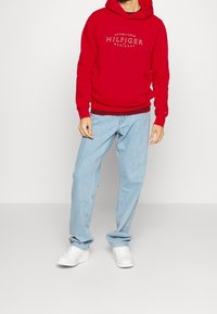 Felpa con cappuccio rossa con logo frontale, abbinata a jeans dritti azzurri e sneaker bianche. L'outfit presenta una vestibilità casual e rilassata.