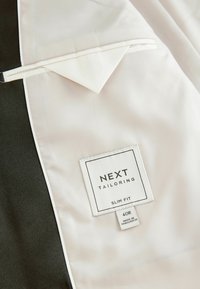 Intérieur d'une veste de costume noire ajustée avec une doublure blanche. Inclut une étiquette d'entretien pour NEXT Tailoring, taille 40R, avec un texte clair et des détails de couture.