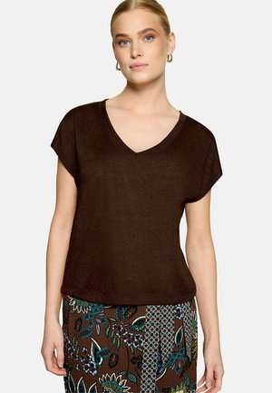 Femme portant un haut marron foncé à manches courtes et col en V, ainsi qu'une jupe marron à motif floral avec des accents bleus et verts.