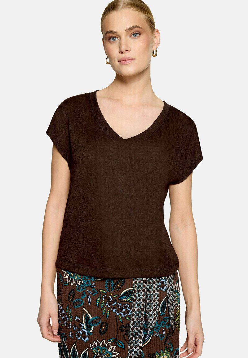 Femme portant un haut marron foncé à manches courtes et col en V, ainsi qu'une jupe marron à motif floral avec des accents bleus et verts.