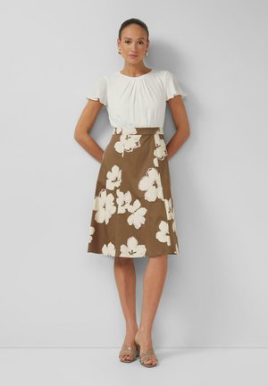 Vrouw draagt een witte blouse met korte mouwen en een bruine A-lijn rok met groot wit bloemenpatroon, staand tegen een effen lichtgrijze achtergrond.