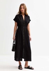 Abito midi in lino nero con maniche corte, chiusura frontale a bottoni e leggera arricciatura in vita. Abbinato a una borsa nera e sandali.