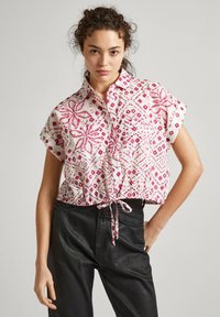 Blusa corta de manga corta con base blanca y patrones florales geométricos en rosa. Atada en la cintura, con cuello de camisa.