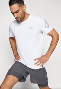 Bílé sportovní tričko se světle modrým logem a třemi šedými pruhy na každém rameni, doplněné o šedé šortky s hladkou texturou.