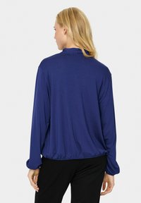 Maglietta blu navy a maniche lunghe con fondo arricciato e texture morbida. La vista posteriore mostra un colletto semplice e una vestibilità ampia. Nessun motivo o decorazione.