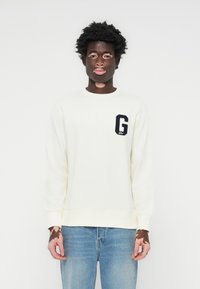 Crèmekleurige sweatshirt met een marineblauwe geborduurde "G"-logo op de linkerborst, gecombineerd met lichtblauwe jeans. Minimalistisch ontwerp en ronde hals.