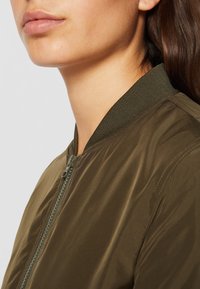 Olive zelená bomber bunda s zipovým uzávěrem, žebrovaným límcem a hladkou texturou. Elegantní, štíhlý design s minimálními akcenty.