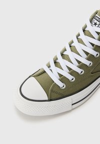 Zöld vászon magasszárú sneaker fehér fűzőkkel, gumis lábujjhéjjal és textúrált talppal. Fekete árnyalatokkal a talp szélén.