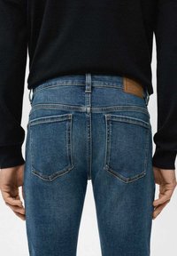 Siniset denimhousut kapealla istuvuudella, takaa nähtynä, joissa on kaksi takataskua ja ruskea nahkainen merkintä vyötäröllä. Yksinkertaiset ompelut.