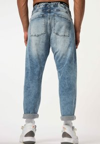 Los jeans de mezclilla azul claro presentan un corte relajado, cinco bolsillos y puños enrollados. La tela muestra una textura deslavada y una cinturilla con marca.