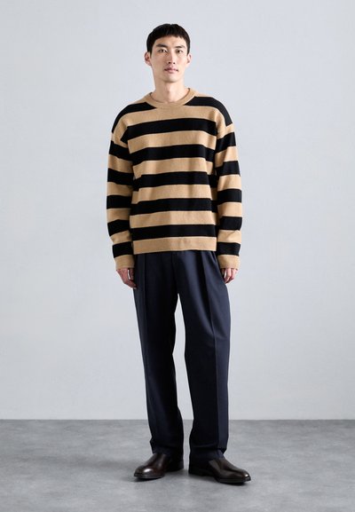 Jersey a rayas en beige y negro, hecho de una suave tela texturizada; corte holgado con cuello redondo, combinado con pantalones formales oscuros y zapatos.