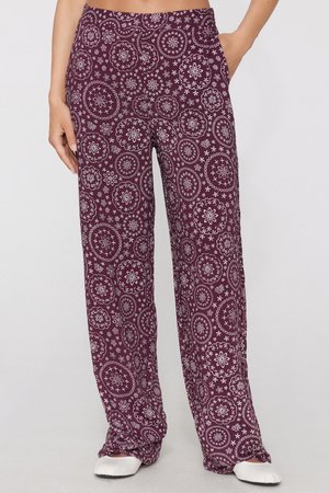 Mujer que lleva pantalones sueltos morados con patrones florales blancos circulares, una mano en el bolsillo, zapatos blancos planos visibles.