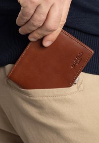 Brown leather wallet partially visible in a beige pocket, featuring smooth texture and a embossed logo on the surface.