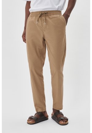 Tan, lightweight, tapered trousers with an elastic waistband and drawstring. Worn with black sandals featuring adjustable straps.