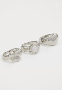 Tres anillos de plata: uno con forma de flor, uno con una piedra opalescente y uno con forma de corazón. Acabado suave, diseños variados.