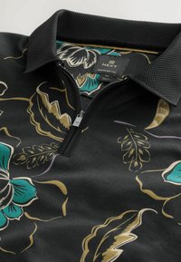 Cămașă polo neagră cu fermoar, cu imprimeu floral în nuanțe de verde, bej și crem, având un guler texturat și eticheta brandului "Next" în interiorul gâtului.