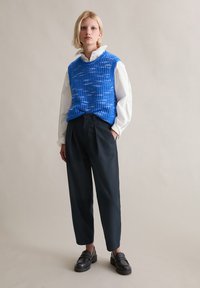 Niebieski bezrękawnik z dzianiny o teksturowanym wzorze, noszony na białej koszuli. Zestawiony z czarnymi zwężanymi spodniami i czarnymi butami wsuwanymi.