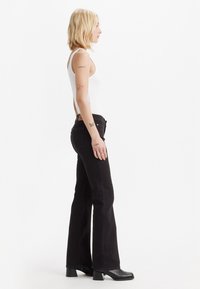 Black flared jeans with a high waistband and five-pocket design, paired with a fitted white tank top. The model stands in profile.
