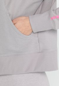 Mano poggiata nella tasca anteriore di un hoodie grigio chiaro con texture, con polsini e orlo a coste, che mostra parte di un logo rosa sulla manica.