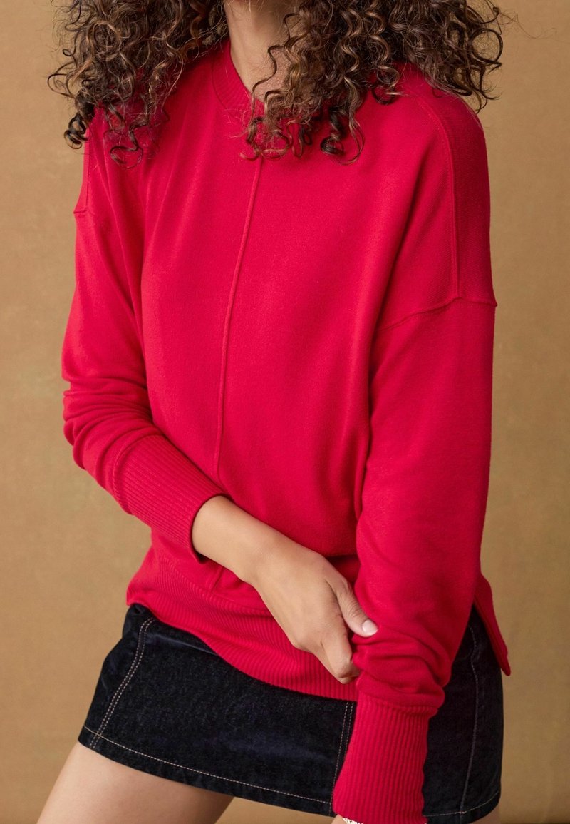 Femme aux cheveux bouclés portant un pull rouge vif à manches longues et une mini-jupe en denim noire sur un fond marron uni.