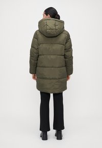 Mujer con el cabello oscuro recogido en un moño, vistiendo una chaqueta de plumas verde oliva con capucha, pantalones negros y botas de tacón negras, de pie de espaldas.