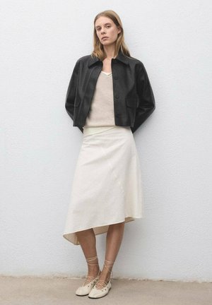 Giacca cropped in pelle nera con colletto rotondo, top in maglia beige, gonna crema asimmetrica e scarpe stringate di colore chiaro.