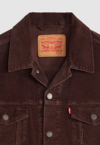Bruine ribfluwelen jasje met een puntige kraag, zilveren knopen en een rode Levi's label. Heeft verticale ribbels en zakken aan de voorkant. Maat M.