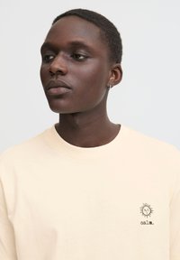 Beige bomulds T-shirt med rund hals, der har et lille solgrafik og ordet "calm." broderet i sort på venstre bryst.