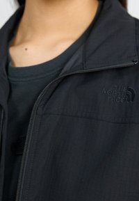 Svart zip-up jacka med en texturerad yta. Har en hög krage och en broderad logotyp på vänster sida. Bärs över en mörk skjorta.